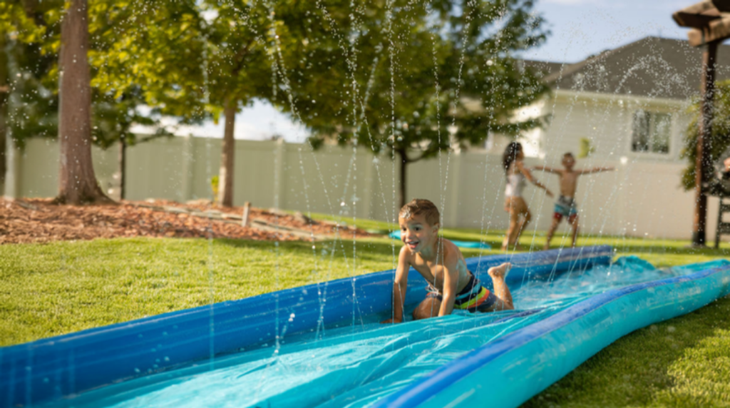 A young boy slides on a blue slip-and-slide in a grassy backyard while two children play in the background on a sunny day.