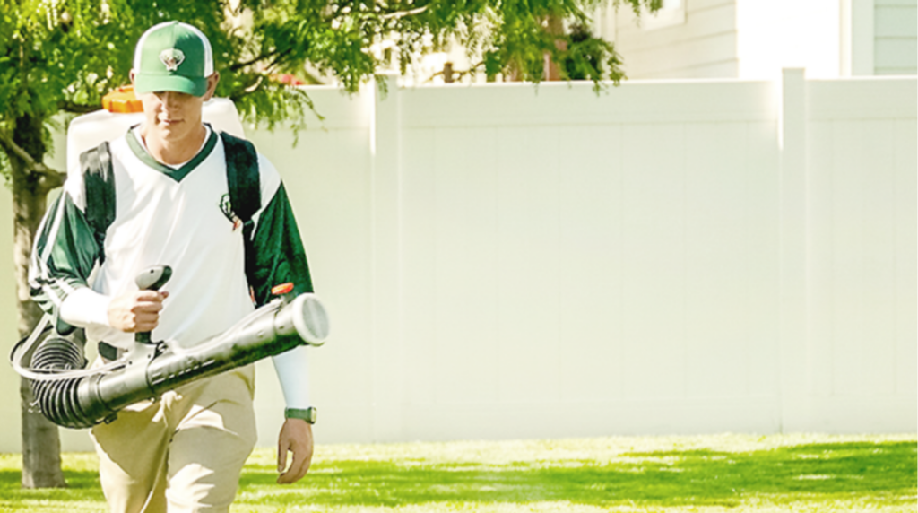 A person wearing a cap and uniform uses a backpack leaf blower on a grassy lawn near a white fence.