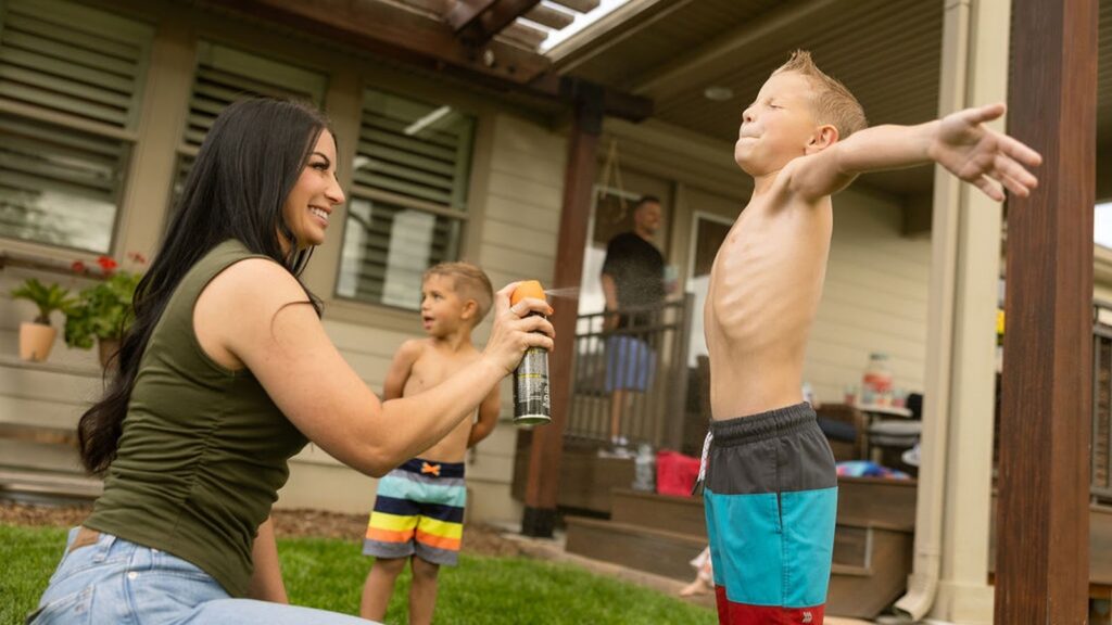 A woman sprays sunscreen or bug spray on a shirtless boy with arms outstretched in a backyard while another child and an adult look on.