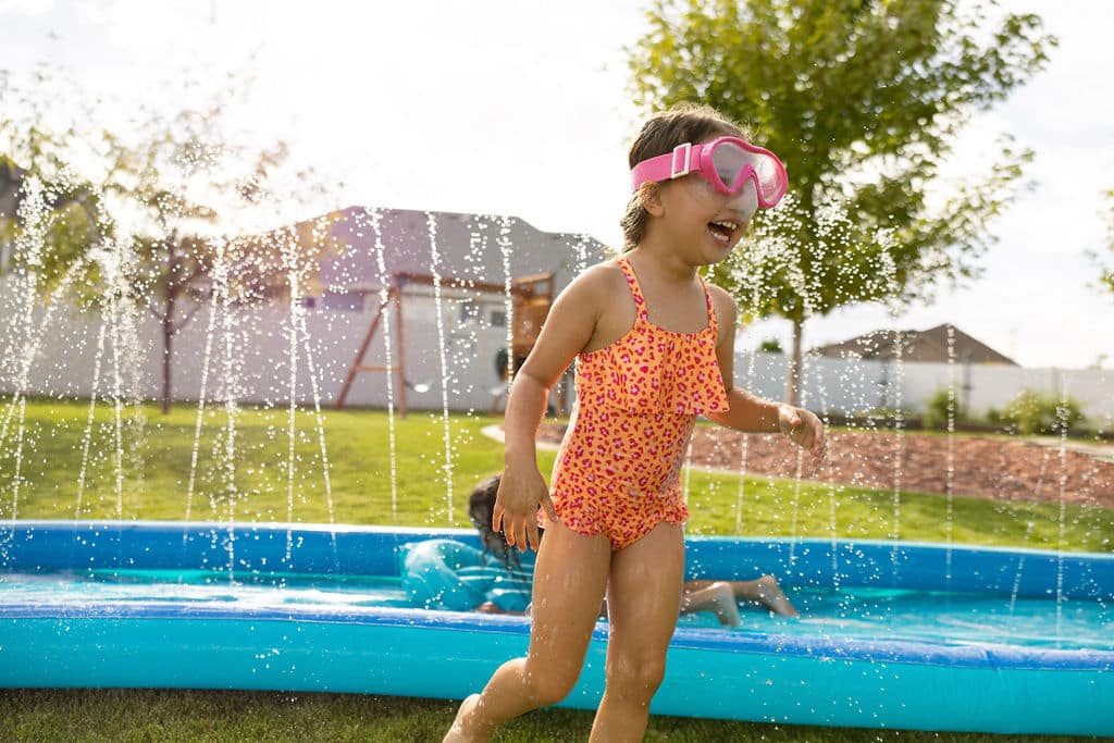 Young child in pink swim goggles and an orange swimsuit runs through water jets next to a blue inflatable pool in a backyard on a sunny day.