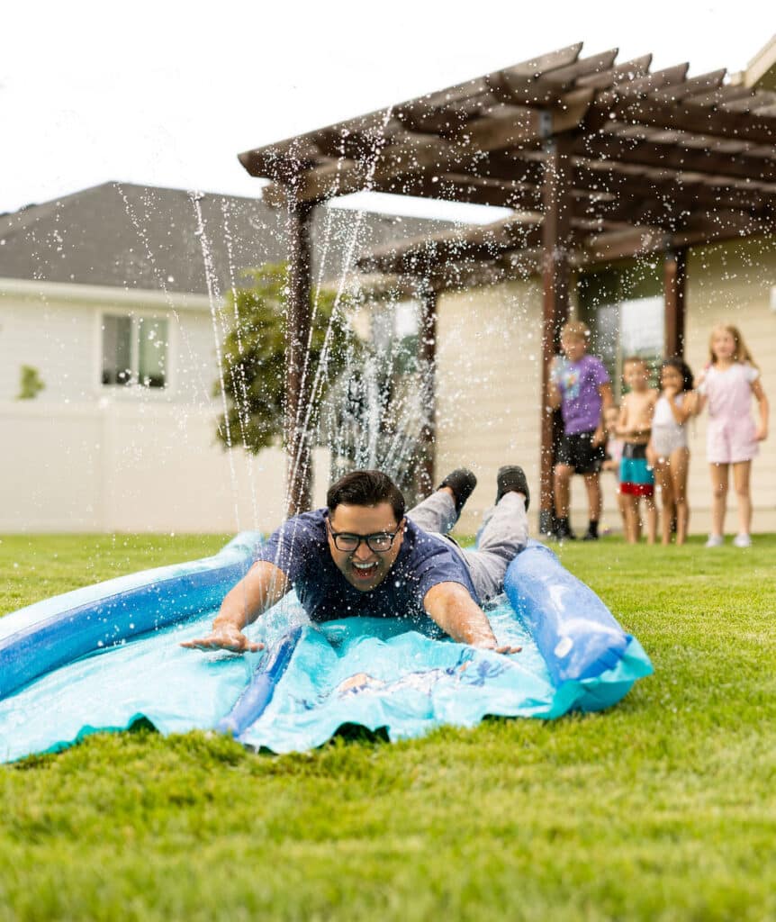 A man slides face-first on a backyard slip and slide while children watch and water sprays upward around him.