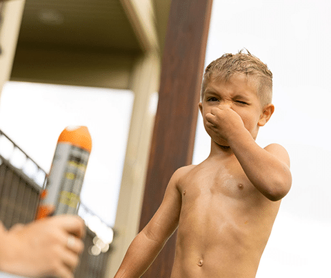 A young boy in swim trunks holds his nose while an adult holds a can of spray sunscreen outdoors in a Utah yard, keeping pets safe with pet-friendly mosquito control.