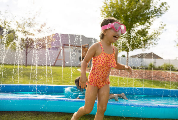 Child wearing pink goggles runs through a backyard sprinkler near a blue inflatable pool, with houses and trees in the background—a classic summer scene in Utah, made even better with proper pest control to keep biting midges at bay.