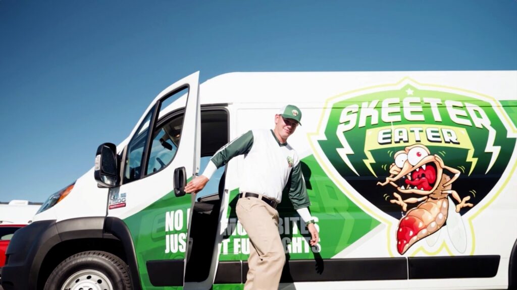 A person in uniform steps out of a van with "Skeeter Eater" and a cartoon mosquito logo on the side under a clear blue sky.