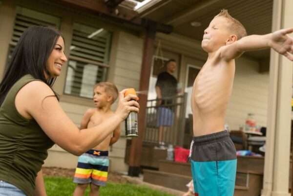 A woman sprays sunscreen on a boy with outstretched arms outside a house, emphasizing the importance of sun safety and mosquito control. Another child and an adult are visible in the background.