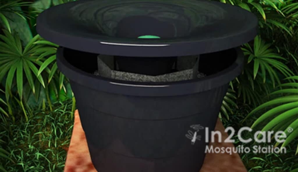 A black In2Care Mosquito Trap sits on a brown surface surrounded by green foliage; the station’s lid is partially open, showcasing advanced mosquito control in action.